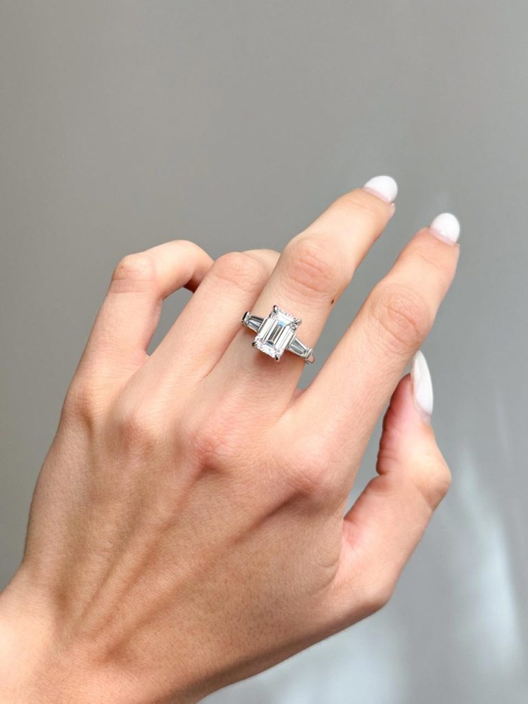 A large clear emerald cut diamond with two tapered baguette cut side stones, set horizontally, on either side, with white pointed prongs holding the stones