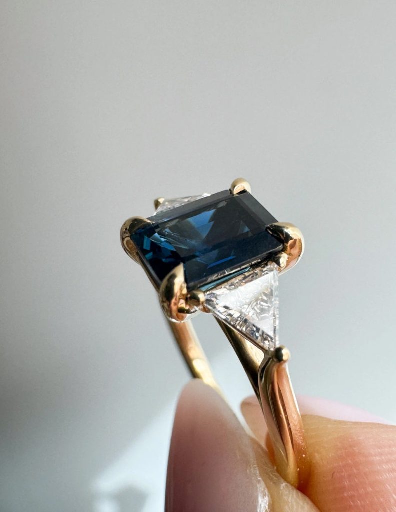 A dark blue emerald cut sapphire, set with four yellow gold prongs, and two triangle diamond accent stones framing its sides